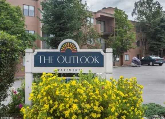 A welcoming entrance sign for Outlook Apartments surrounded by bright yellow flowers, with the apartment building and parking area in the background.