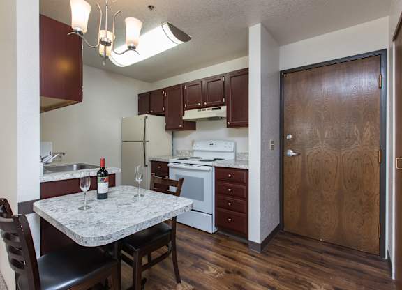 A clean compact kitchen here at Outlook with dark wood cabinets, light countertops, and white appliances, featuring a small dining table under warm overhead lighting, creating a cozy dining and kitchen space.