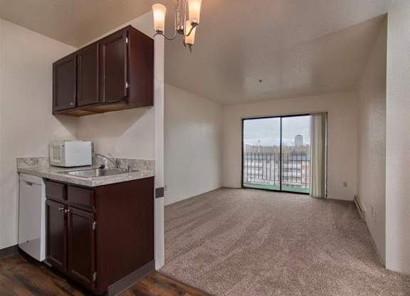 A bright open living space here at Outlook with a compact kitchen featuring dark cabinets, light countertops, and a stainless steel sink, opening into a cozy, carpeted living area with a sliding glass door to a balcony, bringing in natural light.