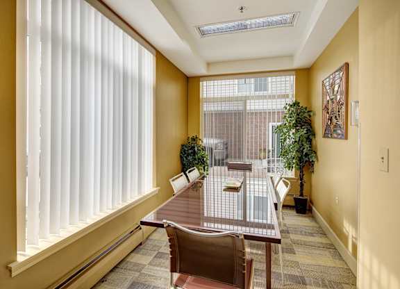 A bright meeting room here at Park Plaza II with a long glass-top conference table and modern chairs, featuring large windows with vertical blinds, warm wall tones, and greenery for a professional, welcoming space.