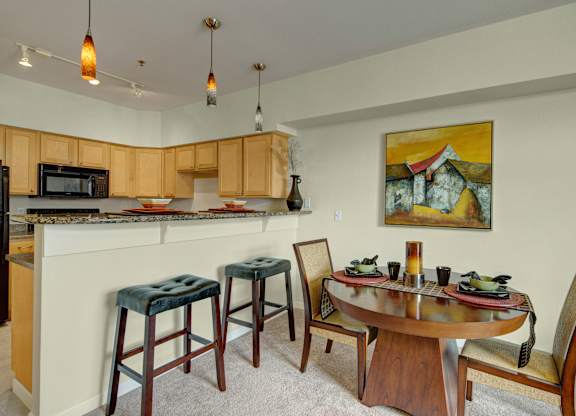 A stylish dining area here at Park Plaza II with a round wooden table and upholstered chairs, featuring a granite breakfast bar with barstool seating, light wood kitchen cabinets, and warm pendant lighting for a modern, open feel.