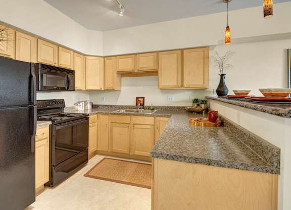 A spacious kitchen here at Park Plaza II with light wood cabinets and speckled granite countertops, featuring a double stainless steel sink, a black stove with built-in microwave, and a dishwasher for a clean, modern setup.