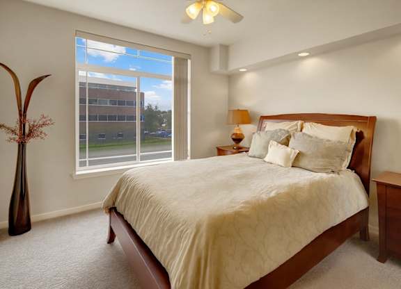 A bright bedroom here at Park Plaza II with plush beige carpeting and a large window with vertical blinds, featuring a cozy bed with neutral bedding, matching wood nightstands, and soft ceiling fan lighting.