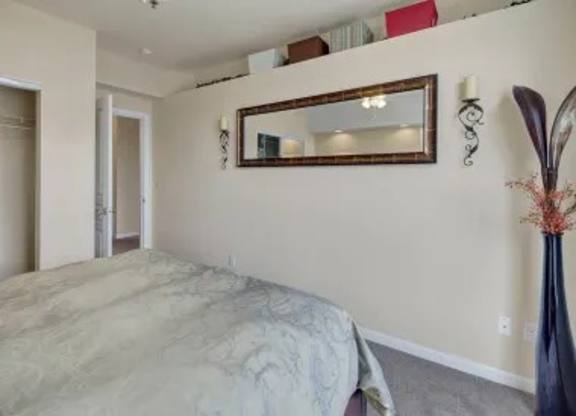 A cozy bedroom here at Park Plaza II with soft neutral carpeting and warm beige walls, featuring a comfortable bed and a large wall mirror that reflects the space for a bright, open feel.