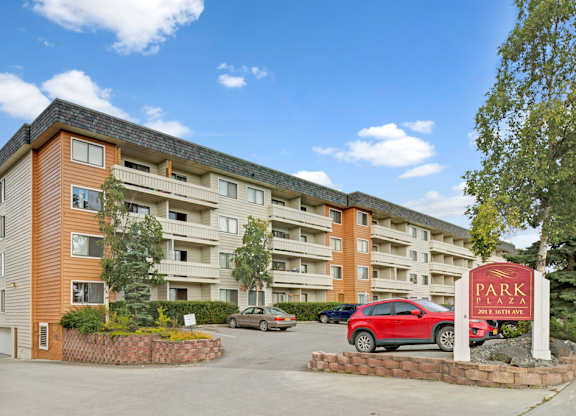 A welcoming exterior view of the Park Plaza apartment community with a multi-story building featuring private balconies, mature landscaping, and convenient on-site parking beneath a bright blue sky.