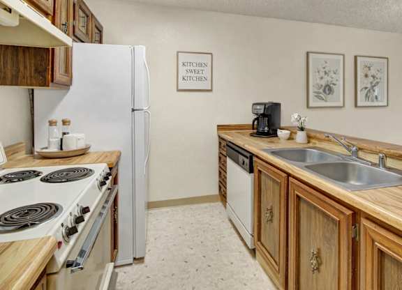 A clean, cozy galley kitchen with warm wood cabinets and butcher-block style countertops, featuring a white refrigerator, white stove, double stainless sink, and simple wall decor with framed botanical prints.