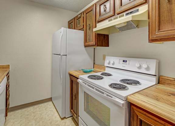 A compact kitchen with warm wood cabinets and light wood-look countertops, featuring a white stove, full-size refrigerator, and built-in dishwasher in a simple, functional layout.