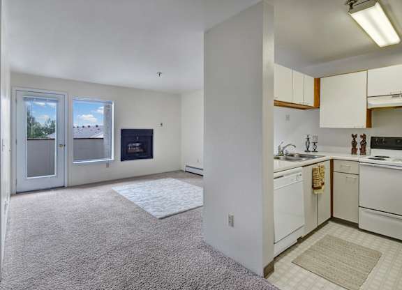 A bright, open-concept living space here at Silver Ridge with soft beige carpet and white walls for a clean, airy feel. A compact kitchen features cream cabinets and white appliances, while the living room includes a cozy fireplace and a glass door leading to a private balcony.