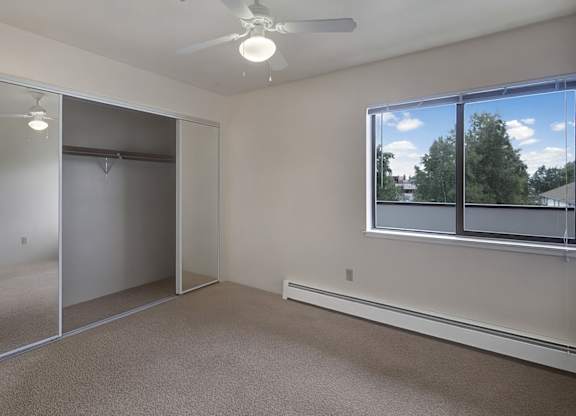 A spacious bedroom here at Silver Ridge with soft beige carpet, light walls, and a bright, airy feel. A large window brings in natural light and a view of greenery, while mirrored closet doors and a ceiling fan with light add comfort and convenience.