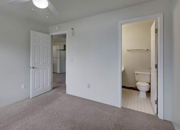 A clean, spacious bedroom here at Silver Ridge with soft beige carpet, light walls, and a bright, simple layout. A ceiling fan with a light adds comfort, while an ensuite bathroom and open doorway leading toward the kitchen create an easy, functional flow.