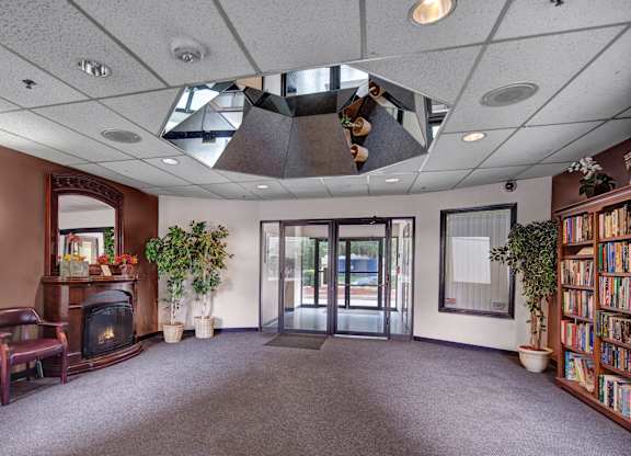 A welcoming lobby here at Silver Ridge with plush carpeting, warm brown accents, and soft overhead lighting for a cozy atmosphere. A decorative fireplace mantle, seating area, and tall greenery create a comfortable entry space, while glass doors and a bookshelf add a polished, community feel.