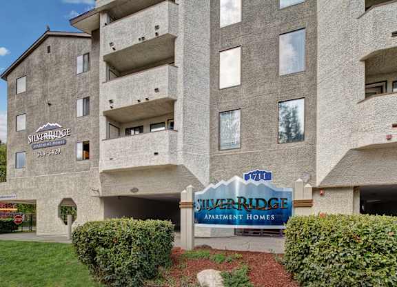 A front exterior view here at Silver Ridge showcasing the community building with private balconies and a clean, modern facade. A Silver Ridge Apartment Homes entrance sign sits in a landscaped area with green grass and shrubs, creating a welcoming first impression.