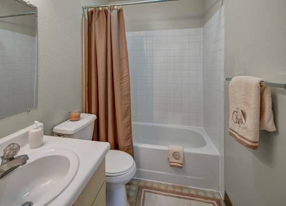 A clean bathroom here at Silver Ridge with a tub-and-shower combo, white tile surround, and a warm neutral shower curtain. A white vanity with a wide countertop sits beside a large mirror, with a toilet and towel bar adding simple everyday comfort.