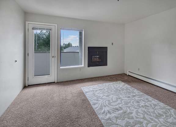 A spacious, empty living room here at Silver Ridge with soft beige carpet and clean white walls for a bright, open feel. A sliding glass door and window bring in natural light, while a built-in fireplace adds a cozy touch and leads out to a private patio or balcony area.