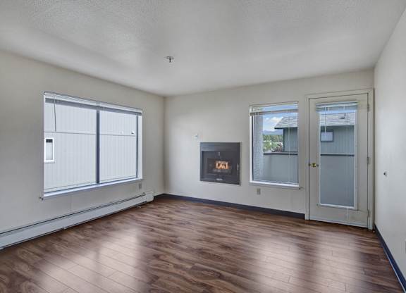A spacious, empty living room here at Silver Ridge with wood-style flooring and soft neutral walls for a clean, open feel. A large window and glass door bring in natural light and lead to a private balcony, while a built-in fireplace adds a cozy focal point.