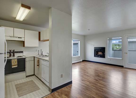 A bright, open living space here at Silver Ridge with wood-style flooring, white walls, and a clean, airy layout. A compact kitchen features cream cabinets and white appliances, while the spacious living room includes large windows, a cozy fireplace, and a glass door leading to a private balcony.