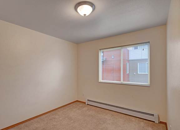 A bright, empty bedroom here at Strawberry Lane with soft beige carpet and warm neutral walls for a clean, cozy feel. A wide window with blinds brings in natural light, creating a simple and comfortable space to make your own.