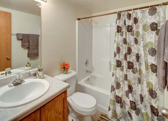 A tidy bathroom here at Strawberry Lane with a bathtub-shower combo, white surround, and a floral shower curtain for a warm touch. A wood vanity with a wide countertop sits beneath a large mirror, with a bright light fixture adding a clean, welcoming feel.