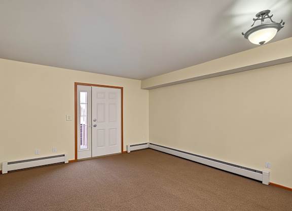 A spacious, empty living room here at Strawberry Lane with soft beige carpet and warm neutral walls for a clean, open feel. A ceiling light brightens the space, while a front entry door with a side window adds natural light and an inviting touch.