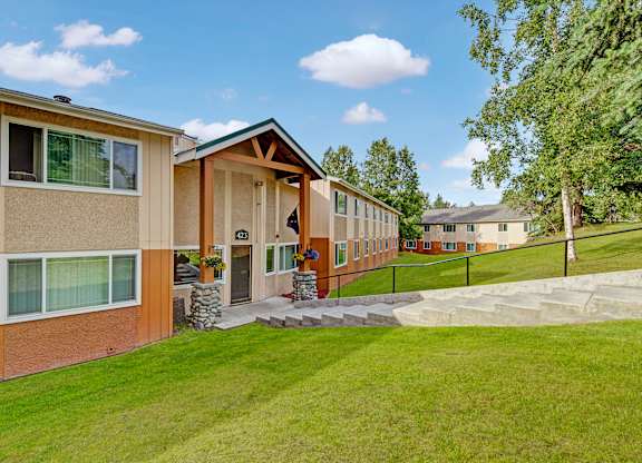 A sunny exterior view here at Taiga featuring a well-kept apartment building with a covered entryway, landscaped greenery, and a gently sloped lawn with walkway access under a bright blue sky.