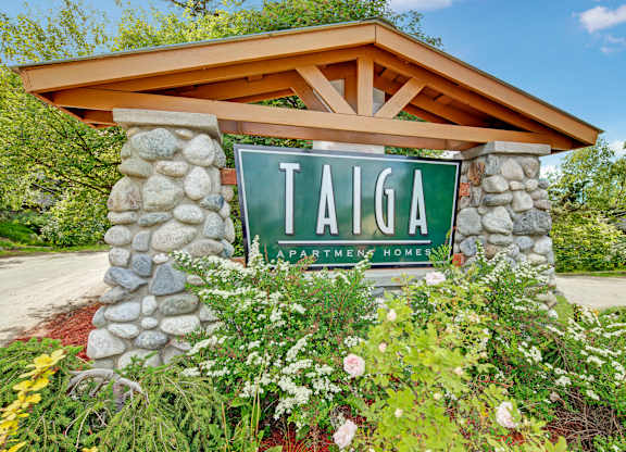 A welcoming entrance sign here at Taiga with a bold green Taiga Apartment Homes display, stone pillars, and lush landscaping that creates a warm, inviting impression.