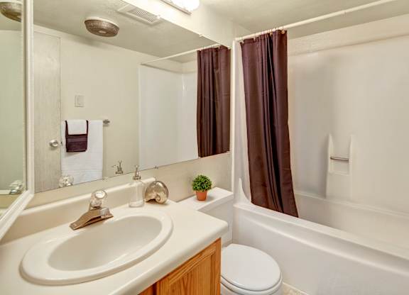 A bright, clean bathroom at Terrace on the Lake featuring a white tub/shower combo with dark brown shower curtains, a large wall mirror, and a spacious vanity with a white countertop and warm wood cabinetry.