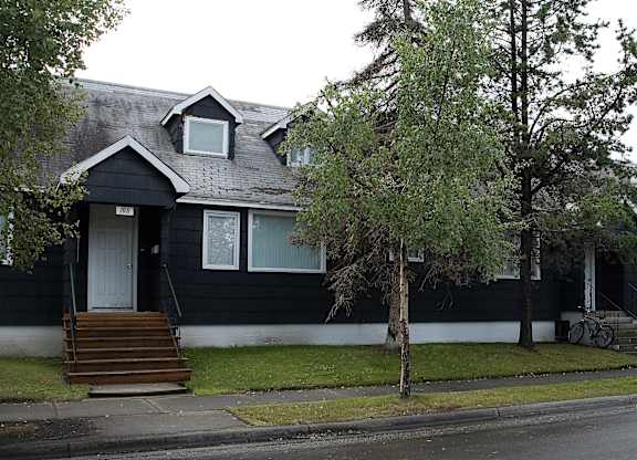 A long single-story building at The Alaskan with dark siding, white trim, and multiple front entrances accessed by small stairways. Mature trees line the sidewalk and grassy lawn, creating a residential neighborhood feel.