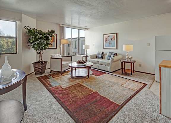 A bright living room at The Alpine featuring a sofa and armchair arranged around a round coffee table on a colorful area rug. Large windows bring in natural light and views of the outdoors. Table lamps, framed artwork, and a potted plant add warmth, with the kitchen area visible nearby.