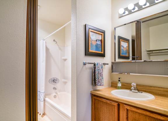 A bright bathroom at The Alpine featuring a single-sink vanity with wood cabinetry and a wide mirror with globe lighting. A tub and shower combo sits behind a white curtain, with built-in shelves for storage. Warm wall art, a hand towel, and simple finishes create a clean and welcoming space.
