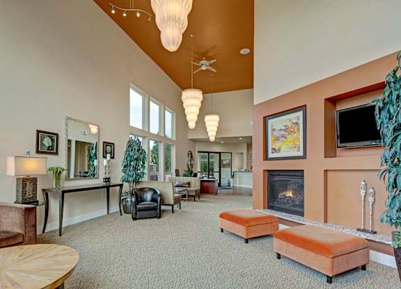 A welcoming lounge at The Alpine with tall ceilings, large windows, and modern hanging lights. Cozy seating includes sofas, armchairs, and upholstered benches near a built-in fireplace. Warm wall tones, framed art, plants, and decorative accents create a comfortable community gathering space.