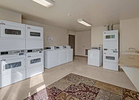 Spacious community laundry room at The Alpine featuring multiple front-loading washers and dryers, including stacked units, a folding counter with sink, and vending-style detergent machine. The bright space has neutral walls, overhead fluorescent lighting, and patterned area rug near the entrance.