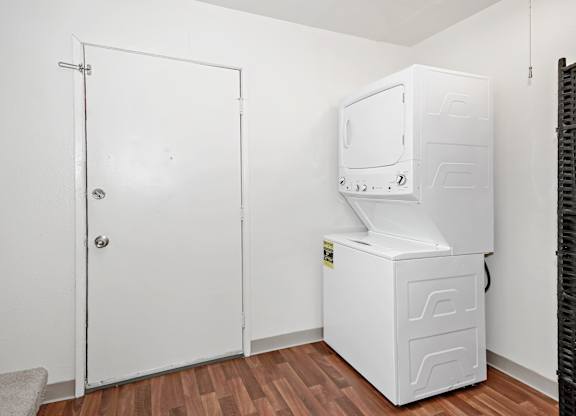 A clean, functional in-home laundry area here at The Glen featuring a stacked white washer and dryer set, durable wood-look flooring, neutral white walls, and a nearby entry door, offering a convenient and efficient space for everyday laundry tasks within the apartment.