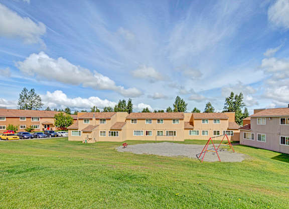 A wide outdoor courtyard here at The Glen featuring well-kept green lawns, a central playground area with red swing sets, surrounding apartment buildings with neutral siding, and open sky views, creating a welcoming shared space for residents to relax, play, and enjoy the landscaped community setting.