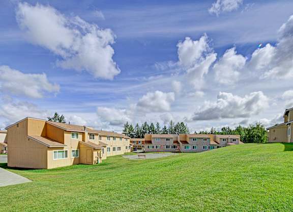 A wide outdoor view here at The Glen featuring tan two-story apartment buildings arranged around an open grassy courtyard, with a gently sloping lawn in the foreground, a small playground area in the center, mature trees lining the property, and a bright blue sky filled with soft white clouds overhead.