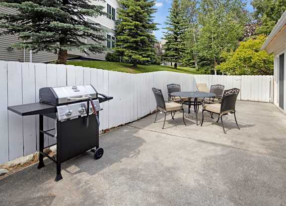 An outdoor amenity space here at The Highlands Luxury, including a barbecue grill, with a rounded outdoor table and chairs, sournded by white fences and concrete flooring.
