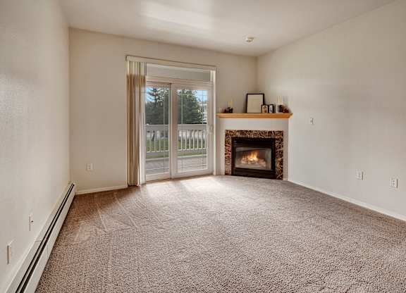 A cozy empty living room here at The Highlands Luxury, with a fireplace against the corner-wall, soft carpeting, and sliding glass door.