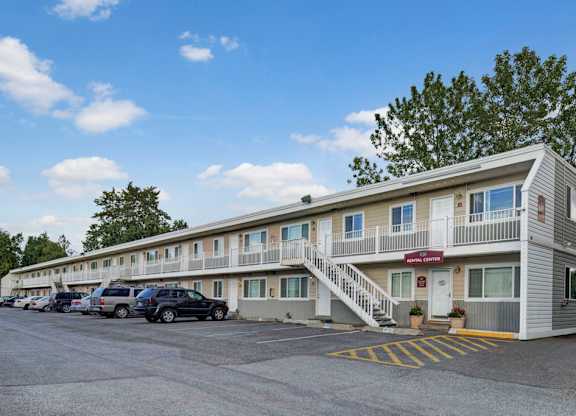 A welcoming exterior view here at The Village at Calais featuring a two-story apartment building with upper-level balconies, a convenient rental center entrance, and a spacious parking area under a bright blue sky.