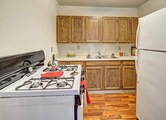 A cozy kitchen at The Village at Calais with wood cabinets, a white stove and refrigerator, a double sink, and wood-look flooring, accented by small colorful dishes and towels on the counters.