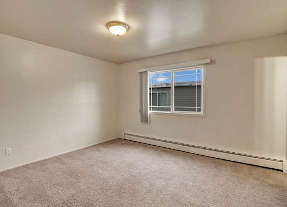 A spacious, empty bedroom here at The Village at Calais with soft beige carpet, clean white walls, and a wide window with vertical blinds that lets in natural light.