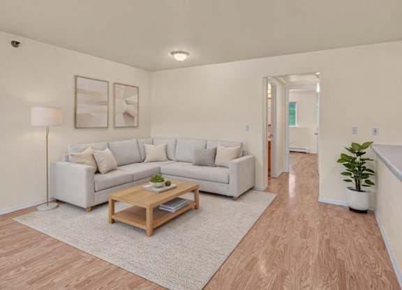 A bright living room here at Timber Ridge with light wood flooring, a cozy light-gray sectional sofa with neutral throw pillows, and a wooden coffee table set on a soft area rug, creating a clean and modern feel.