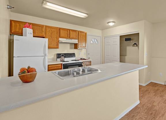 A spacious kitchen here at Timber Ridge with warm wood cabinets, light countertops, and a double stainless steel sink along a wide breakfast bar, featuring white appliances and an open layout with a nearby closet for extra storage.
