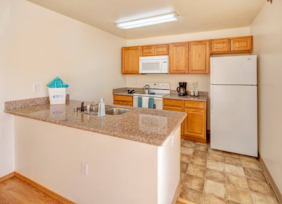A bright kitchen here at Timber Ridge with warm wood cabinets, speckled granite-style countertops, and a stainless steel sink built into a large breakfast bar, featuring white appliances and a clean, open layout with tiled flooring.