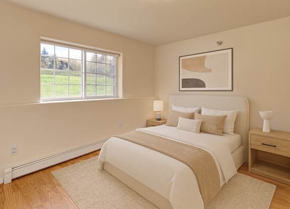 A bright bedroom here at Timber Ridge with light wood flooring, soft neutral bedding, and a cozy bed framed by matching nightstands, accented by modern wall art and natural light from a large window with a peaceful outdoor view.