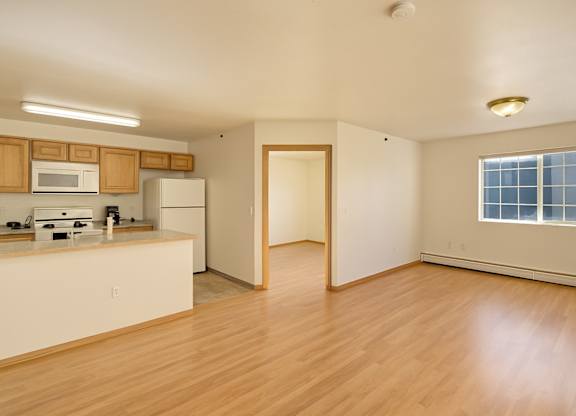 A spacious, open living area here at Timber Ridge with light wood flooring, a large window for natural light, and an open kitchen featuring warm wood cabinets, light countertops, and white appliances for a bright, inviting layout.