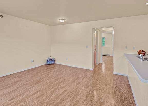 A spacious, empty living area here at Timber Ridge with light wood-style flooring, soft neutral walls, and an open layout leading into the kitchen with a long countertop and stainless steel sink, creating a bright and flexible space for living and dining.