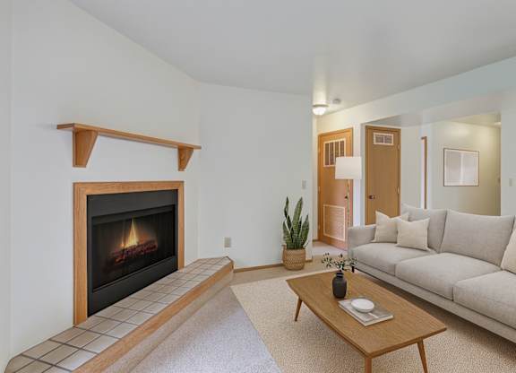 A cozy living room here at Wildwood Estates with a warm corner fireplace, soft neutral carpeting, and a light beige sofa paired with a simple wooden coffee table and tasteful greenery for a clean, inviting feel.