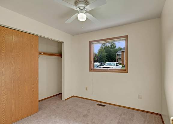 A bright, empty bedroom here at Wildwood Estates with soft carpet, a ceiling fan with overhead light, and a spacious closet with sliding doors, plus a window overlooking the community.