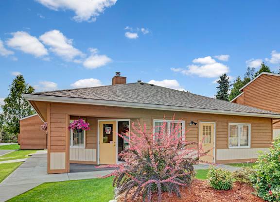 A charming exterior view here at Wildwood Estates featuring the community office building with well-kept landscaping, bright green lawns, and colorful flowers under a sunny blue sky.