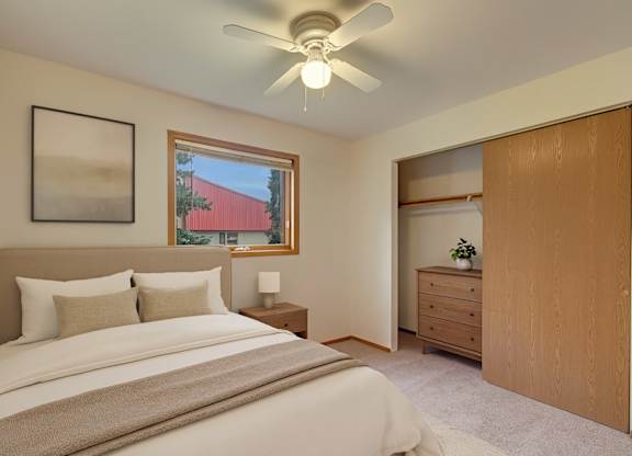 A relaxing bedroom here at Wildwood Estates with soft carpet, a ceiling fan with overhead light, and a cozy bed with neutral bedding, plus a window for natural light and a spacious closet with sliding doors.