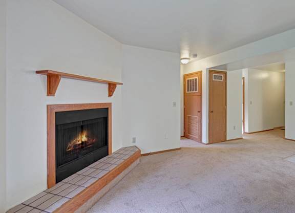 A spacious, empty living room here at Wildwood Estates with soft neutral carpet, a cozy corner fireplace with a tile surround, and an open layout that flows into the adjacent kitchen area.